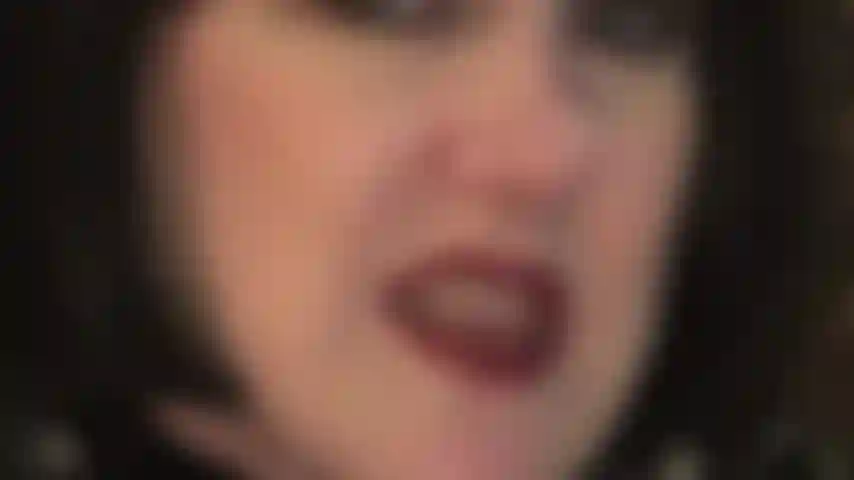 Rostro de cerca con peluca bob negra, provocando con labios pintados de rojo y lengua mojada, así como dientes y manos enguantadas de cuero que disfrutarían aferrándose a tus bolas.